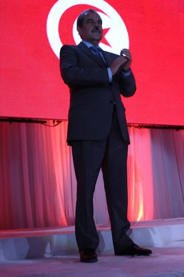 Mondher Zenaidi standing on a stage in front of a giant screen showing the Tunisian flag
